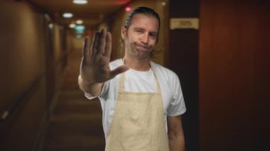 Beyaz tişörtlü ve önlüklü sarışın bir adam otel koridorunda avuç içini açıyor, sınır uyarısını reddediyor..
