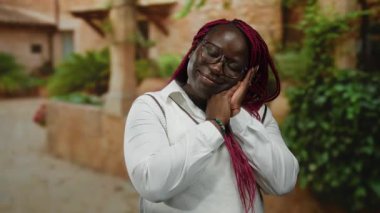 Kırmızı örgülü ve gözlüklü kadınlar şehir sokaklarında tuğla binalar ve yeşilliklerle uyur. Açık bir şehir ortamında sakinliğini ve rahatlığını ifade eder..