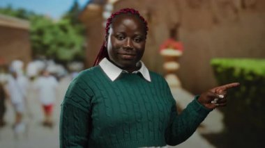 Arka planda bulanık yayalar ve yeşilliklerle dolup taşan bir caddede dikilirken yeşil süveter içinde gülümseyen bir kadın..