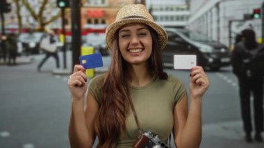 Elinde şapka, kahverengi saç, giysi, genç, İspanyol, şehir, gündüz, araba, arka plan ile dolup taşan caddede elinde kredi kartı tutan kadın gülümsüyor.