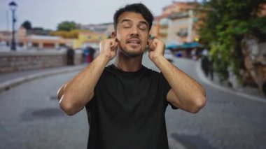 Genç İspanyol adam şehir sokaklarında kulaklarını tıkıyor canlı binalar ve dinamik atmosferi ve çeşitli kültürel ortamları vurgulayan bulanık arka planlı şehir manzarası sergiliyor..