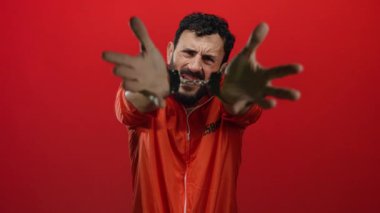 Portakal rengi hapishane tulumlu sakallı, elleri kelepçeli İspanyol bir adam, canlı kırmızı bir arka plana karşı duruyor, hapis ve kısıtlama sahnesini betimliyor..