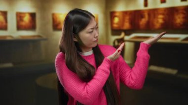 Bir sanat galerisinde pembe kazak giyen bir kadın, sıcak ışıklandırmalı bir iç müze dekorunu sergileyen resimlere karşı düşünceli bir ifade sergiliyor..