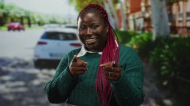 Kırmızı saçlı bir kadın şehir dışındaki bir sokak sahnesinde parmaklarıyla gülümsüyor, şehir arka planı güneşli bir günde arabaları park ettiğini ve hareket ettiğini gösteriyor, neşe ve güveni vurguluyor..