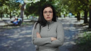 Güneşli yeşil bir park ortamında kolları çaprazlanmış, gözlüklü ve gri kazaklı, düşünceli ve şaşkın bir ruh halini canlandıran bir kadın..