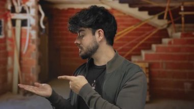 İnşaat alanının içinde bitmemiş tuğla duvarlar ve görünen kablolarla avucunun içini gösteren sakallı İspanyol bir adam..