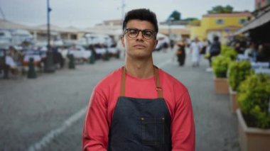 Gözlüklü ve önlüklü genç adam şehir caddesinde açık hava kafeleri ve canlı bir atmosferi olan güvenli bir şekilde duruyor..