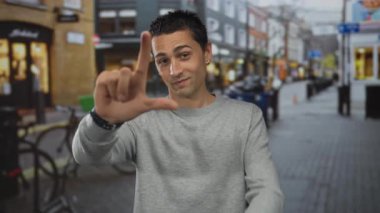 Genç İspanyol adam elinde beceriksiz bir jest yapıyor ve kalabalık bir şehir sokağında kamerayı işaret ediyor..