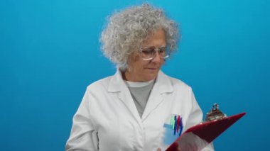 Kadın bilim adamı gri saç, laboratuvar önlüğü ve güvenlik gözlüğü takarak mavi duvara izole edilmiş bir panoyu inceliyor. Laboratuvar ortamında odak ve üst düzey uzmanlık gösteriyor..