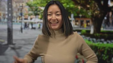 Arka planda ağaçlarla dolu canlı bir şehir sokağında barış işareti yapan genç bir kadın gülümsüyor ve mutluluk ve pozitiflik yayıyor..