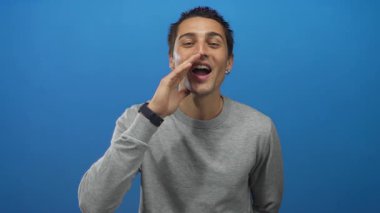 Katı mavi arka planda gri süveter giyen İspanyol genç adam neşeli bir ifadeyle coşkuyla bağırıyor gibi görünüyor..