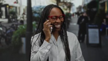 Kentsel sokak ortamında akıllı telefonla konuşan, neşeli bir ifadeyle gülümseyen, gözlük takan, şehir ortamında iletişim ve mutluluk gösteren bir kadın..