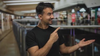 Bir alışveriş merkezinde gülümseyen, siyah tişörtlü İspanyol bir adam, evin içinde parlak perakende mağazaları ve canlı atmosferle çevrili bir heyecan olduğunu gösteriyor..