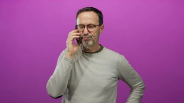 Akıllı telefondan konuşan olgun bir adam canlı pembe bir arka plana karşı izole edilmiş, gözlüklü ve sıradan bir süveter giymiş, sohbete dalmış..