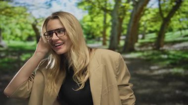 Parkta gülümseyen bir kadın, sarı saçlı, gözlüklü, yeşil ve güneş ışığıyla çevrili bej ceketli rahat bir açık hava yaratıyor..