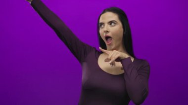 Canlı mor duvara karşı heyecanla işaret eden, zarif kıyafetler giyen, neşe ve coşkuyu ifade eden bir kadın..