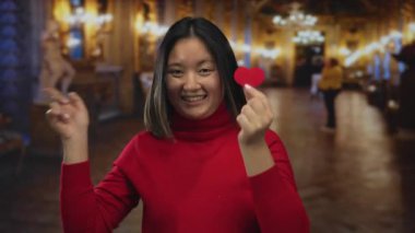 Kapalı alanda kırmızı kalp kesiği tutan bir kadın müzenin içini gösterirken gülümsüyor ve sıcak, davetkar bir atmosferi var..