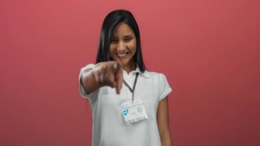 Gülümseyen ve el kol hareketi yapan bir kadın canlı kırmızı arka planda beyaz polo ve isim etiketi takarak dışardaki neşeli yüz ifadesiyle pozitifliğini ve duygularını ifade ediyor..