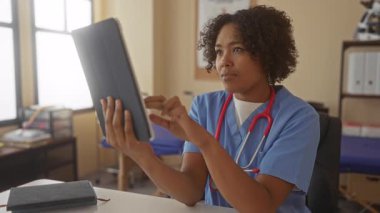 Mavi önlük giyen kadın doktor güneş ışığı binasında kırmızı steteskoplu tablet tutuyor; sakin profesyonel odaklanma.