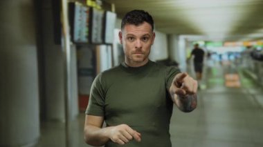 Havaalanı terminalinde dikilirken işaret eden dövmeli genç bir adam yoğun bir iç mekan ortamında yön değiştiriyor ve odaklanıyor..