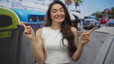 İspanyol genç kadın, işlek şehir caddesinde geri dönüşüm konteynırını işaret ediyor; keşif merakı.