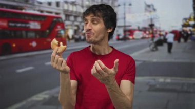 Kırmızı gömlekli genç adam ısırılmış bir donutu kemirerek düşünceli bir bakışla şehir caddesine bakıyor..