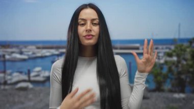 Genç İspanyol bir kadın arka planda teknelerle deniz kenarında jest yapıyor, sakin bir sahil ortamının açık hava sahnesini gösteriyor..