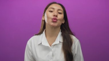 Beyaz tişört giyen genç bir kadın kalp hareketi yapıyor ve mor stüdyoda öpücük yolluyor..