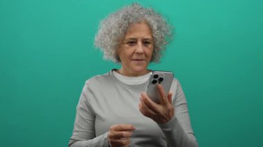 Gri saçlı kadın yeşil arka plana karşı akıllı telefon kullanıyor, izole modern ortamda neşe ve coşku ifade ediyor..