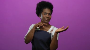 Kadın gülümsüyor ve açık avucuna işaret ediyor, önlük giymiş, pembe bir arka planın karşısında duruyor, bir hoş geldin ya da reklam jestini canlandırıyor..