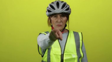 Sarı arka planda güvenlik yeleği ve miğfer takan kıdemli İspanyol kadın, kararlılıkla işaret ediyor ve işaret ediyor, tecrit edilmiş bir ortamda güvenlik ve iletişim kuruyor..