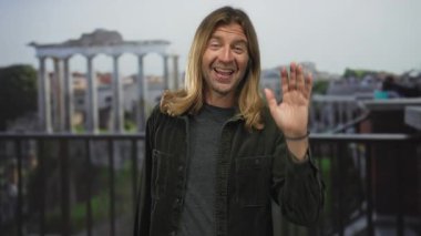 Uzun sarı saçlı ve gülümseyen yüzlü bir adam Roma 'daki harabelerin önünde el sallıyor..