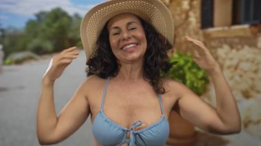 İspanyol kadın bikinili ve şapkalı eski bir kasabada güneşli bir günün tadını çıkarıyor. Dışarıdan neşe ve rahatlama yayıyor..