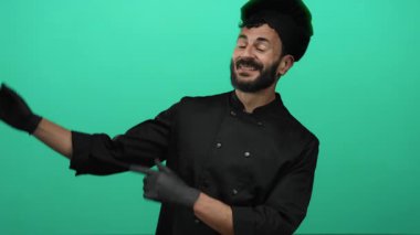 Sakallı İspanyol erkek aşçı canlı yeşil bir fon karşısında şakacı bir şekilde yan yatmış, siyah şef kıyafeti ve uyumlu bir şapka giymiş, aşçılık hevesini somutlaştırıyor..