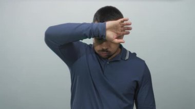 Adam alnından ter siler ve parlak ışık altında açık gri stüdyoda elleri ile gözleri korur; rahatlama.