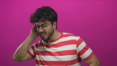 Sakallı, gözlüklü, çizgili gömlek giyen genç İspanyol adam elini kulağına dayayıp canlı pembe bir arka planda dikkatlice dinliyor..