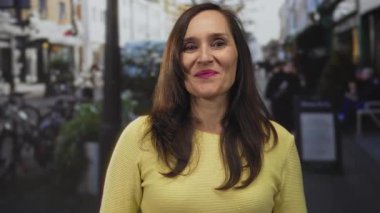 İşlek bir cadde terasında gülümseyen, sarı kazak giyen, iyi bir jest yapan, bulanık kentsel geçmişi olan, Avrupalı bir kafe atmosferi öneren bir kadın..