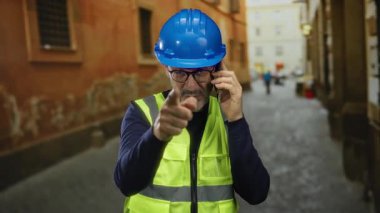 Mavi miğferli ve sarı yelekli bir adam, şehir sokaklarında telefonla konuşuyor, gülümsüyor ve işaret ediyor, orta yaşlı inşaat işçisi dışarıda..