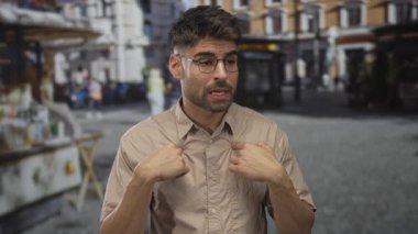 Gözlüklü ve bej gömlekli bir adam, parke şehir sokağında dükkan önleri olan bir caddede parmak göğsü işaret ediyor..