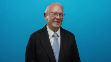 Takım elbiseli yaşlı bir adam parlak mavi bir arka planda dikilirken göz kırpıyor, açık mavi kravat ve gözlüğünü vurguluyor, neşeli bir iş davranışını çağrıştırıyor..