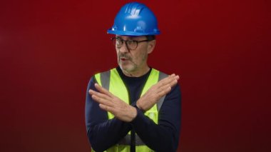 Güvenlik yeleği ve miğferli adam kırmızı arka planda çapraz kollara sahip orta yaşlı, İspanyol erkek tarafından izole edilmiş duvarda gösterilen güvenlik endişesini gösteriyor..