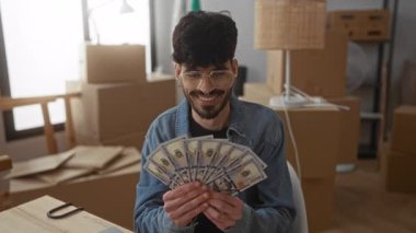 Oturma odasında gülümseyen genç bir adam yeni evdeki kutuların arasında oturarak bize dolarları tutuyor. Taşınırken finansal başarı öneriyor..