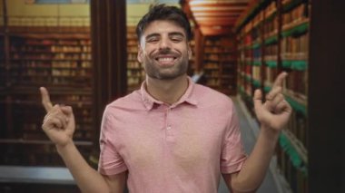 Pembe gömlekli genç adam gülümsüyor ve binadaki ahşap kütüphane rafını işaret ediyor..