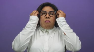 Gözlüklü kadın, mor arka planda ellerini kaldırmış, şok halinde. Beyaz bir gömlek giyiyor. Duygularını ve şaşkınlığını canlı bir arkaplanda aktarıyor..