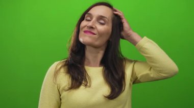 Parlak yeşil bir arkaplan, uzun koyu renk saçlar ve sarı bir kazak giyen neşeli bir yüz ifadesine güvenen bir kadın..