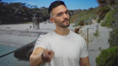 Deniz ve doğa ile çevrili, ciddi bir ifadeyle kameraya bakan genç İspanyol bir adam, açık havada günlük beyaz bir gömlek giyiyor..