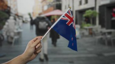 Kalabalık bir şehir caddesinde elinde yeni Zelanda bayrağı tutan bir adam. Arka planda bulanık insanlar ve binalar var. Ulusal gurur ve kültürel çeşitlilik sergiliyor..