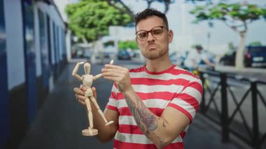 Dövmeli bir adam, şehir arka planında tahta bir model tutuyor. Gözlük ve çizgili gömlek giyiyor..