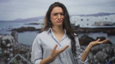 Uzun siyah saçlı, omuz silkmiş elleri ve deniz kenarındaki bir bina tarafından çizilmiş kaşları olan genç bir kadın..