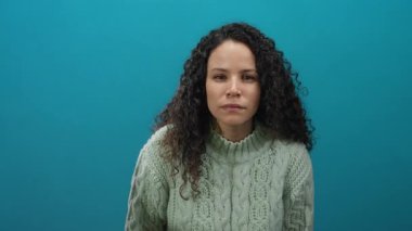 Kazaklı, kıvırcık saçlı bir kadın izole edilmiş mavi duvara karşı duruyor. Düşünceli ve anlamlı görünüyor. Basit ama canlı bir ortamda bir dizi duygu taşıyor..